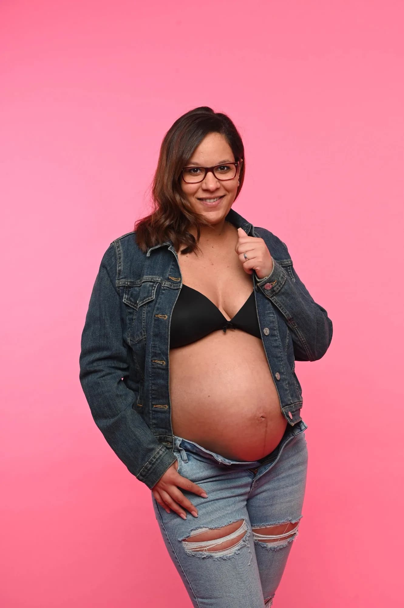 Lachende Schwangere in Jeansjacke und Jeans zeigt ihren Babybauch vor pinkem Studiohintergrund